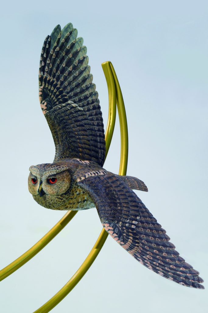 オオコノハズクのバードカービング作品。真鍮パイプでできた月をバックに、悠々と飛翔しています。